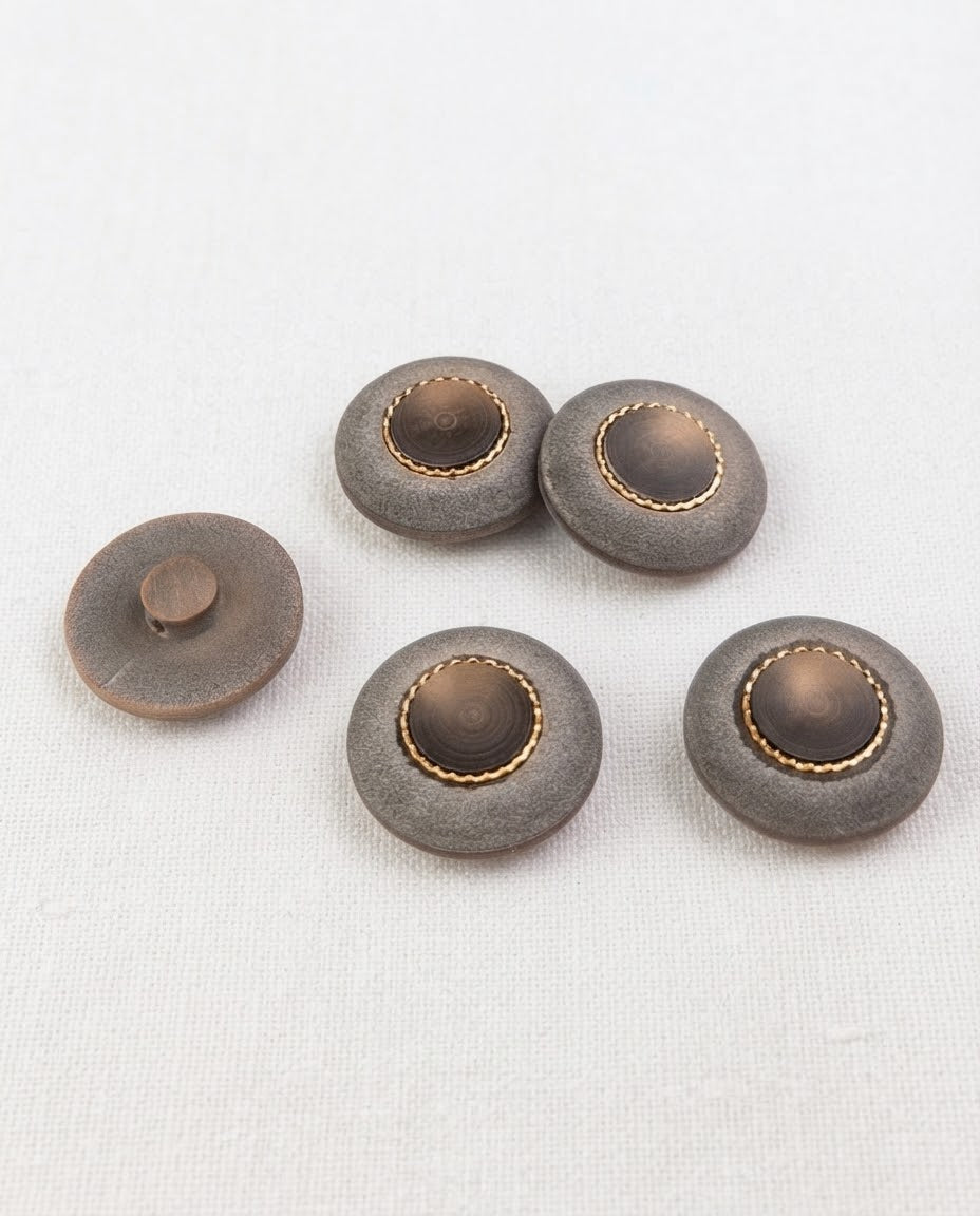 Boutons ronds gris mat et doré de 22 mm vus de biais sur une toile écrue, dont un retourné montrant sa queue