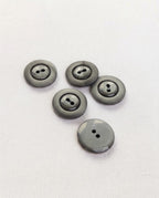Boutons ronds gris foncé de 17 mm à 2 trous vus de biais sur un tissu blanc, dont un vu de dos