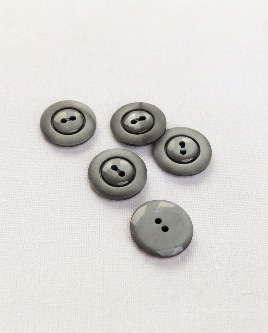 Boutons ronds gris foncé de 17 mm à 2 trous vus de biais sur un tissu blanc, dont un vu de dos