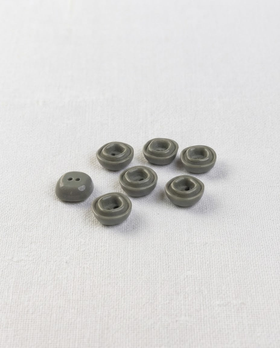 Petits boutons carrés gris vert de 12 mm vus de biais, relief carré autour des deux trous, sur un tissu blanc