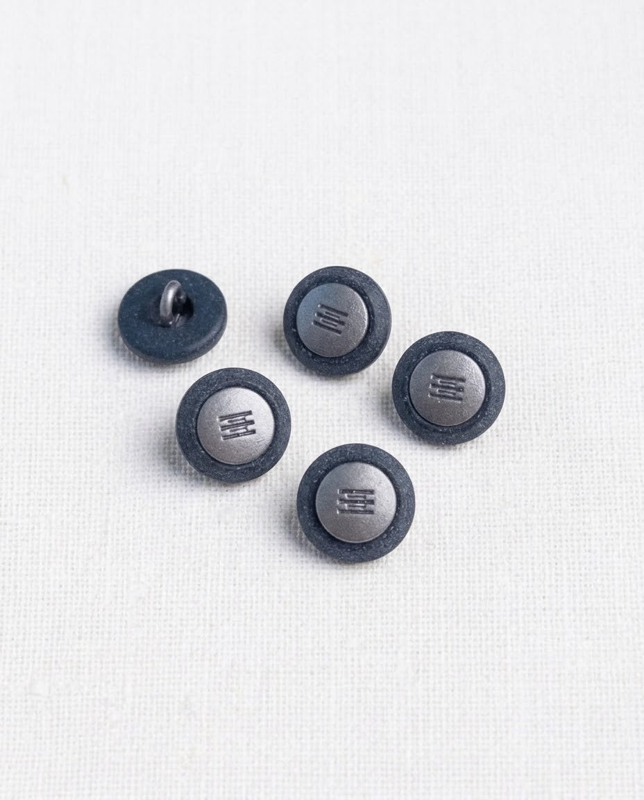 Boutons bleu jean à centre argenté de 14 mm vus de dessus sur un tissu blanc, avec un bouton retourné pour voir sa queue