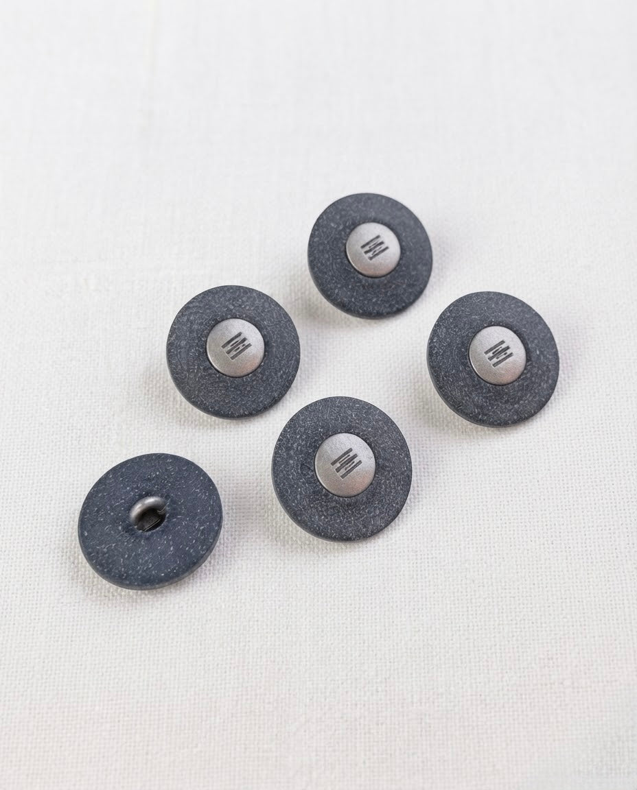 Boutons bleu jean à centre argenté de 22 mm posés sur un tissu blanc, avec un bouton retourné qui montre la queue