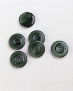 Six boutons vert sapin de 22 mm à 2 trous posés sur une toile écrue, marbrures plus sombres