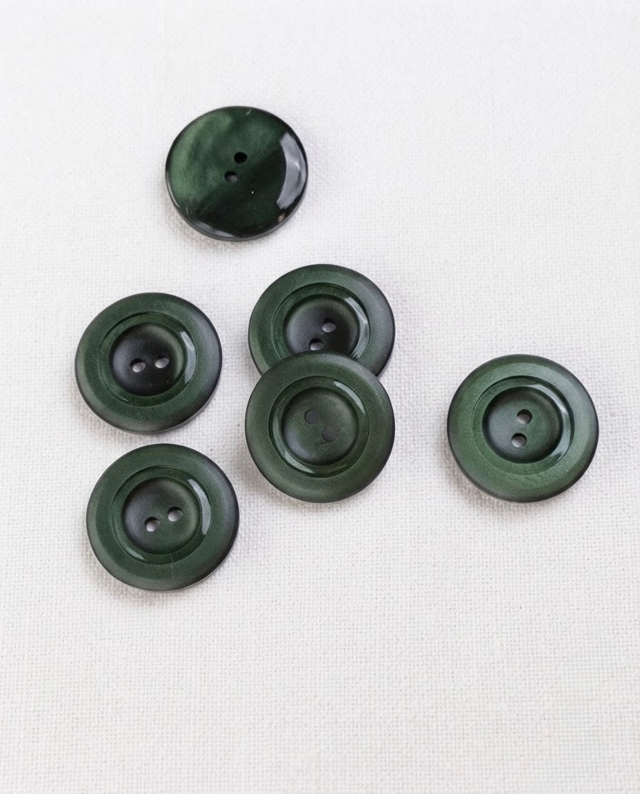 Six boutons vert sapin de 22 mm à 2 trous posés sur une toile écrue, marbrures plus sombres
