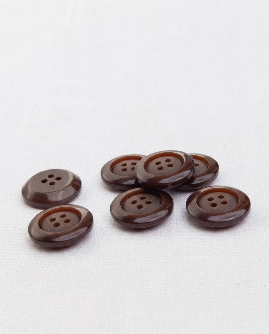 Sept boutons marron chocolat de 17 mm à 4 trous posés sur une toile blanche, dont un retourné