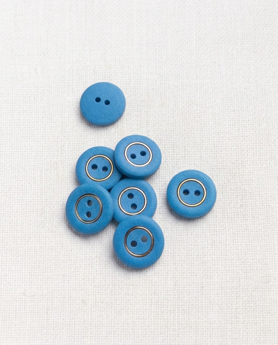 Boutons ronds bleus à cercle doré de 14 mm à 2 trous posés sur un tissu blanc