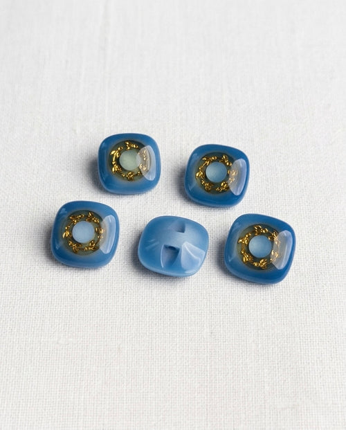 Cinq boutons carrés aux angles arrondis de 16 mm, bleu brillant avec une couronne dorée autour d'un centre bleu clair, dont un vu de dos avec la queue encastrée