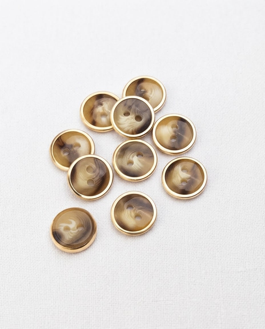 Dix petits boutons ronds effet corne cerclés de doré, 12 mm, 2 trous, vus de dessus sur une toile blanche, dont un retourné