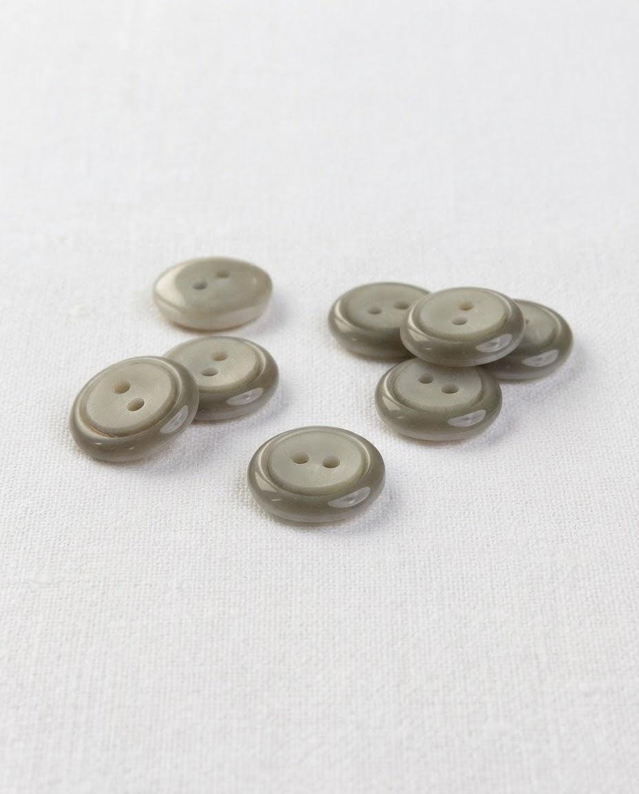 Boutons ronds gris vert brillants de 13 mm à 2 trous posés sur une toile blanche, vus de biais