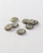 Boutons ronds gris vert brillants de 13 mm à 2 trous posés sur une toile blanche, vus de biais