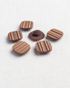 Boutons carrés marron rainurés de 16 mm posés sur un tissu blanc, un bouton retourné montre sa queue