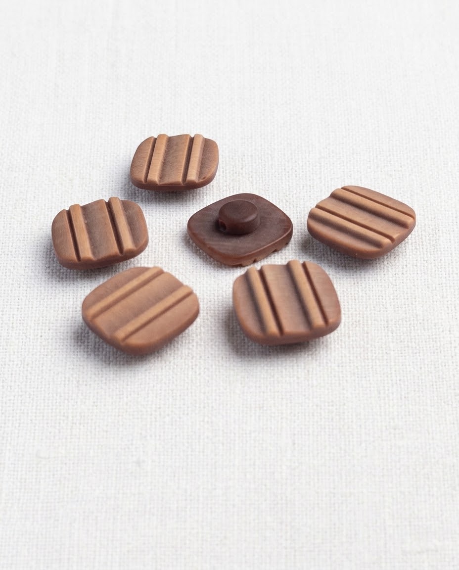 Boutons carrés marron rainurés de 16 mm posés sur un tissu blanc, un bouton retourné montre sa queue