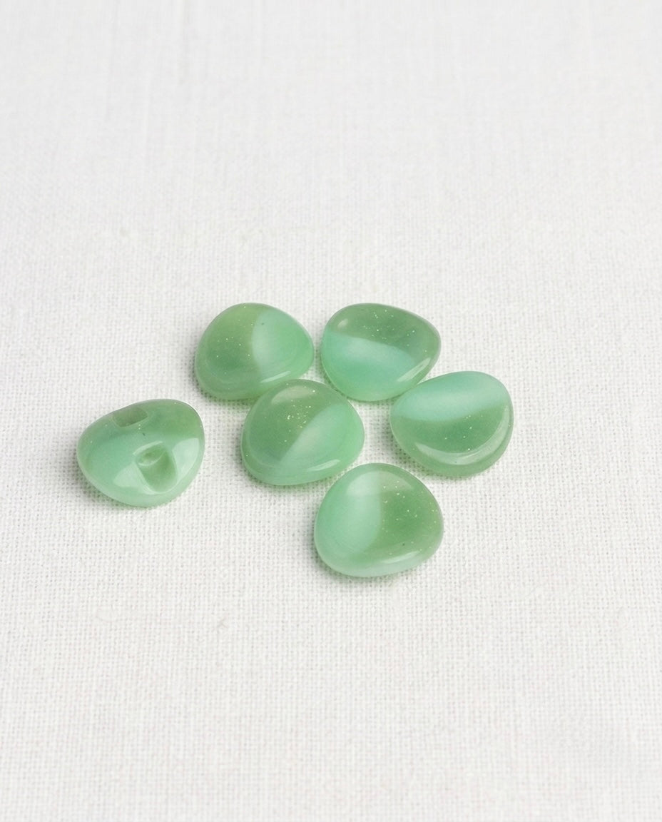 Boutons vert amande au bord ondulé de 15 mm posés sur une toile écrue, reflets marbrés