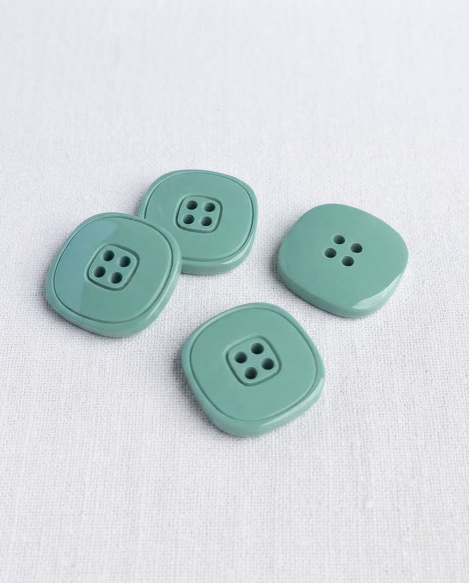 Quatre boutons carrés vert de gris de 20 mm à 4 trous vus de dessus, dont un sans cadre gravé visible