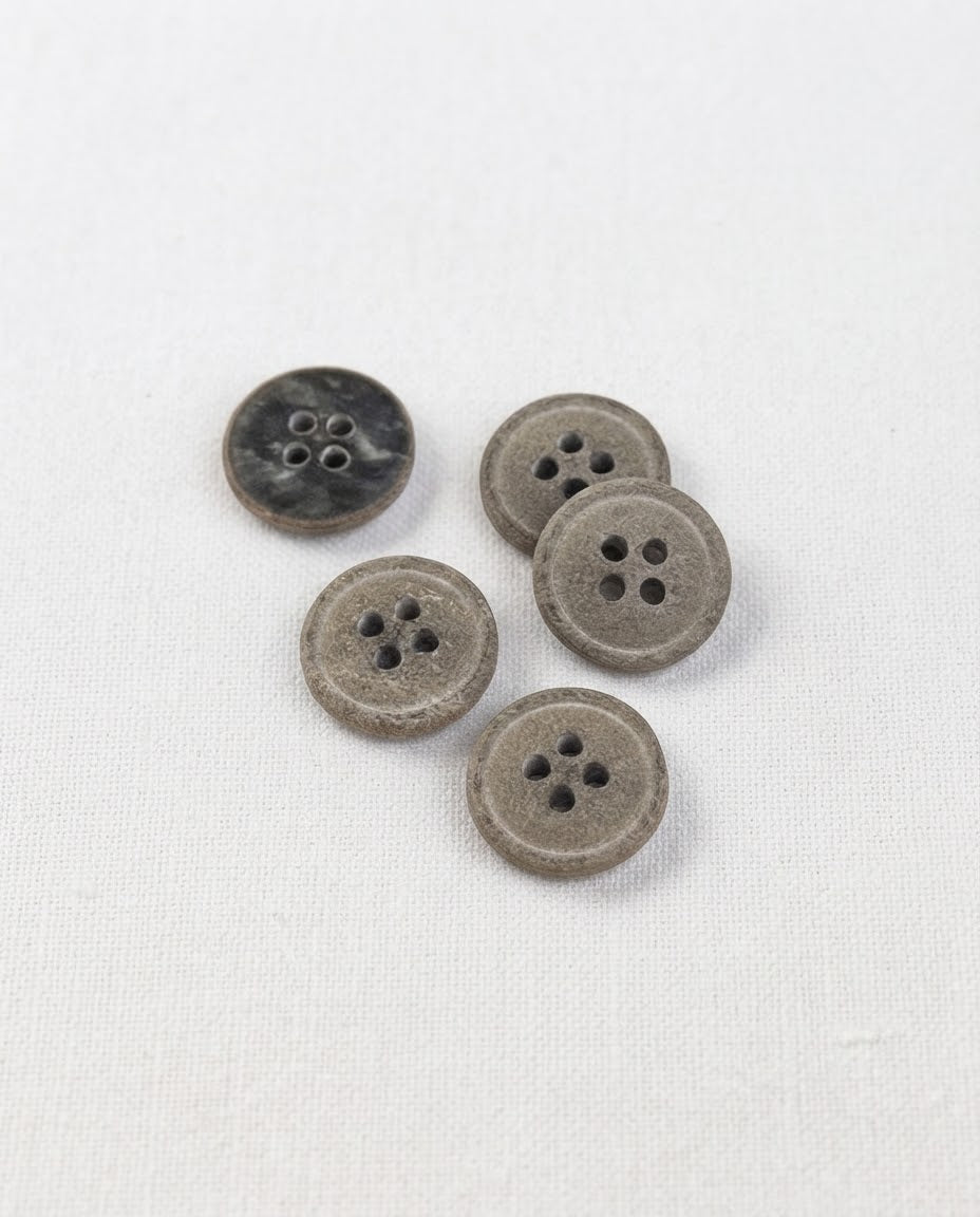 Boutons ronds gris kaki mouchetés de 15 mm à 4 trous vus de dessus sur un tissu blanc, dont un retourné
