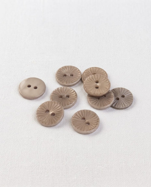 Boutons ronds beige taupe de 15 mm à 2 trous posés sur un tissu blanc, avec leur motif soleil gravé en relief