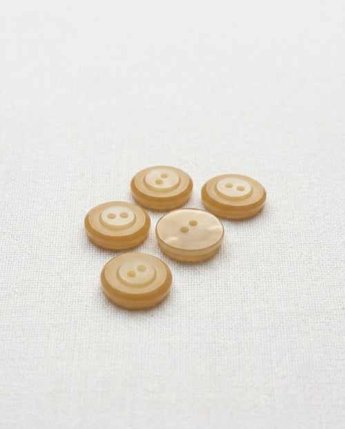 Boutons ronds beige de 14 mm à 2 trous vus de biais sur un tissu blanc, montrant leur rebord à deux tons