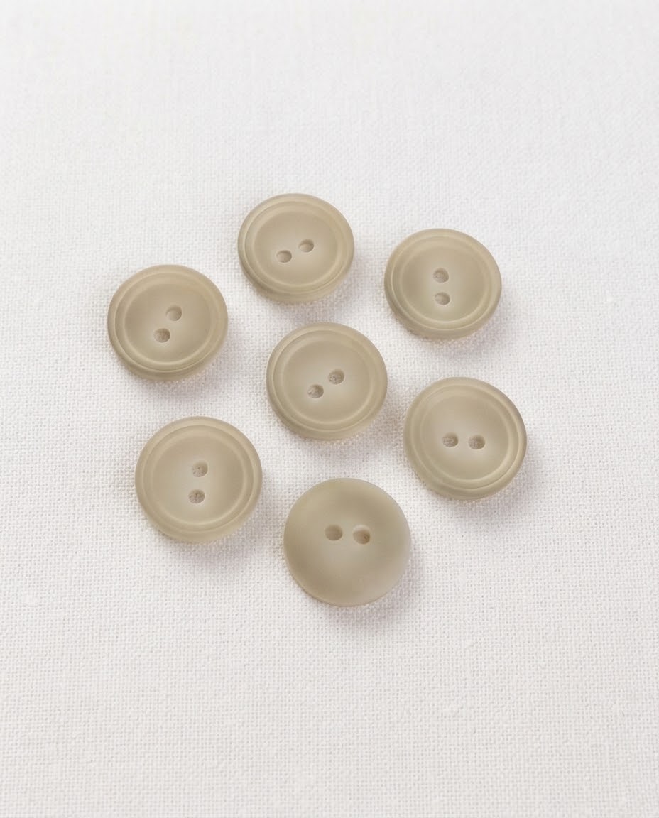 Boutons ronds effet corne beige grisé de 15 mm à 2 trous vus de dessus sur un tissu blanc, dont un retourné