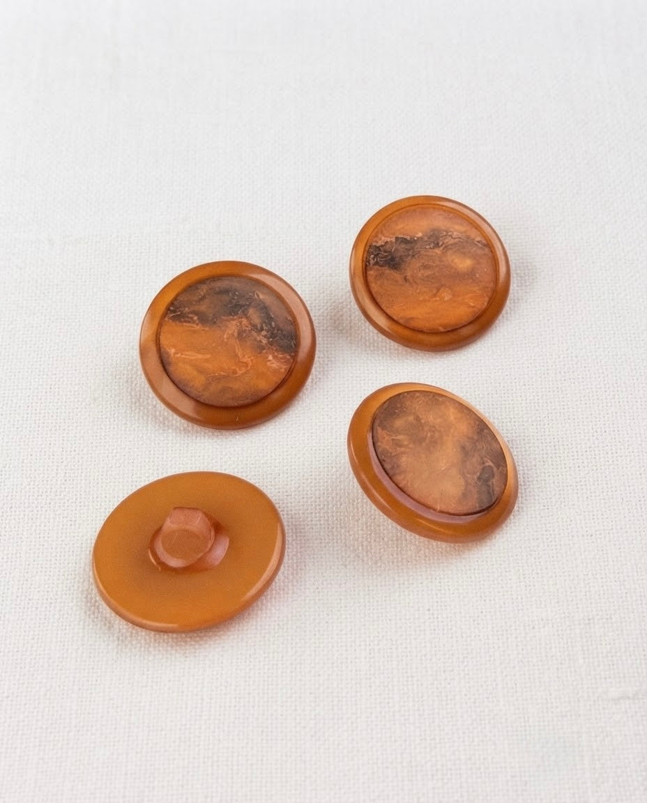 Boutons cuivre marbrés de 22 mm vus de dessus sur un tissu blanc, un bouton retourné montrant la queue