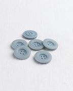 Boutons ronds bleu gris mat de 17 mm à 4 trous vus de biais sur un tissu blanc