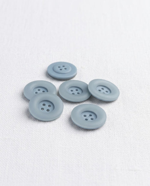 Boutons ronds bleu gris mat de 17 mm à 4 trous vus de biais sur un tissu blanc