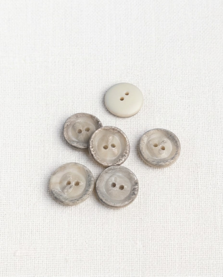 Boutons ronds crème à bord marbré gris de 16 mm vus de dessus sur un tissu blanc, un bouton retourné uni