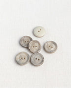 Boutons ronds crème à bord marbré gris de 16 mm vus de dessus sur un tissu blanc, un bouton retourné uni