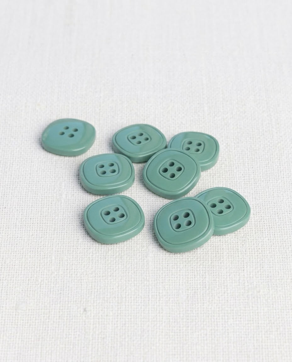 Boutons carrés vert de gris de 17 mm à 4 trous posés sur une toile écrue, dont un vu de dos