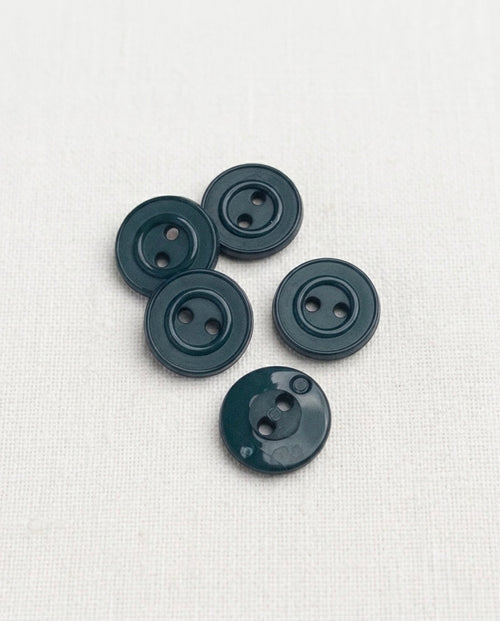 Boutons bleu pétrole de 15 mm à 2 trous posés sur un tissu blanc, un bouton retourné montrant le dos lisse