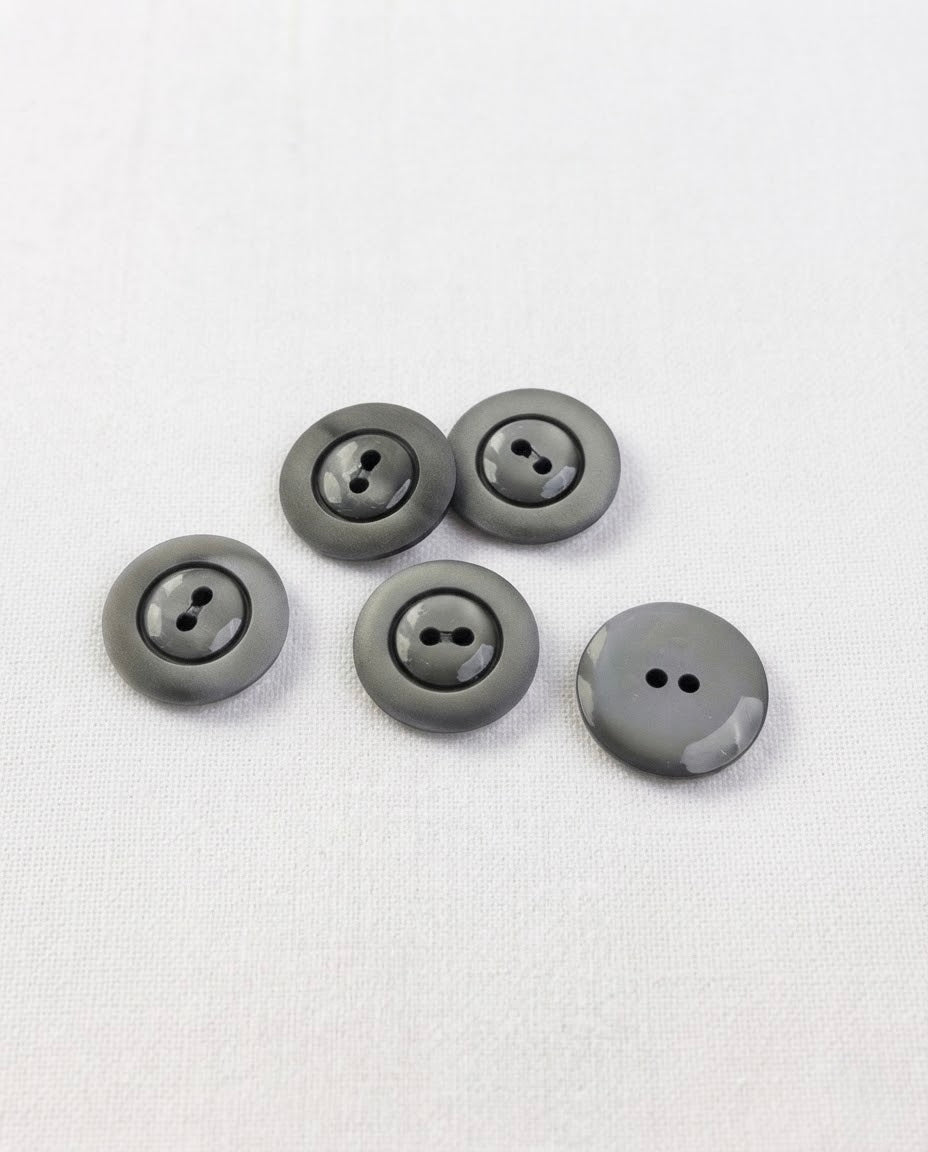 Boutons ronds gris ardoise de 17 mm à 2 trous posés sur une toile écrue, bord mat et centre creusé brillant