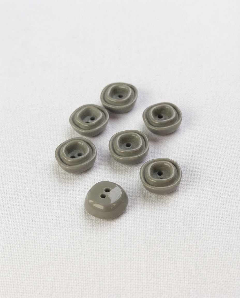 Sept petits boutons carrés gris vert brillants de 12 mm à 2 trous, avec un bourrelet carré en relief, dont un vu de dos