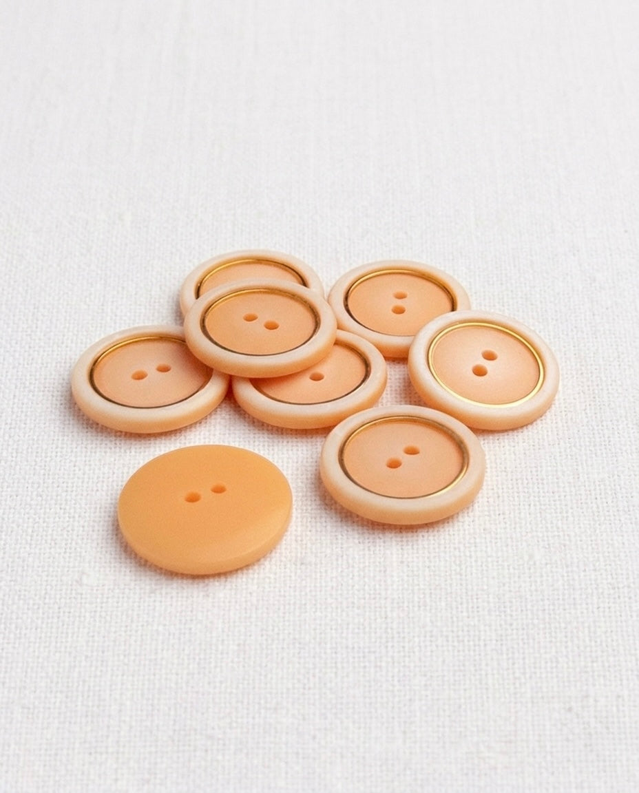 Sept boutons ronds de 18 mm à 2 trous, centre pêche mat, bord crème et fin filet doré, dont un vu de dos pêche uni